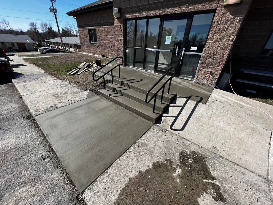 Exterior view of a building with a newly constructed concrete ramp and steps leading to glass doors, with black handrails.