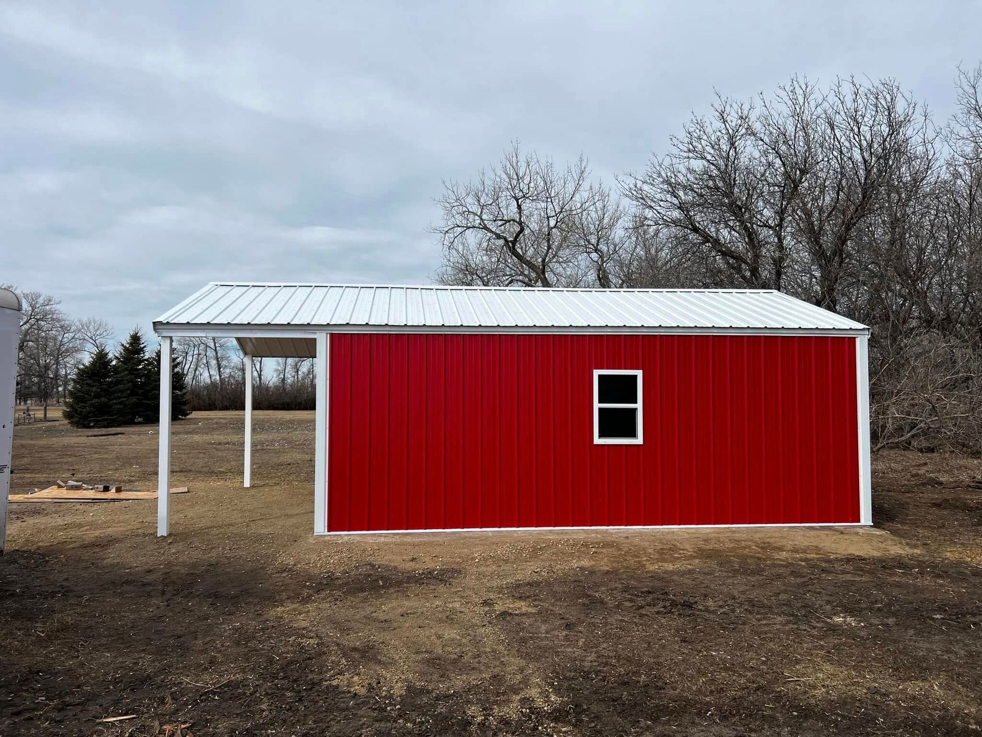 Red metal shed with white trim and a covered carport, under a cloudy sky.