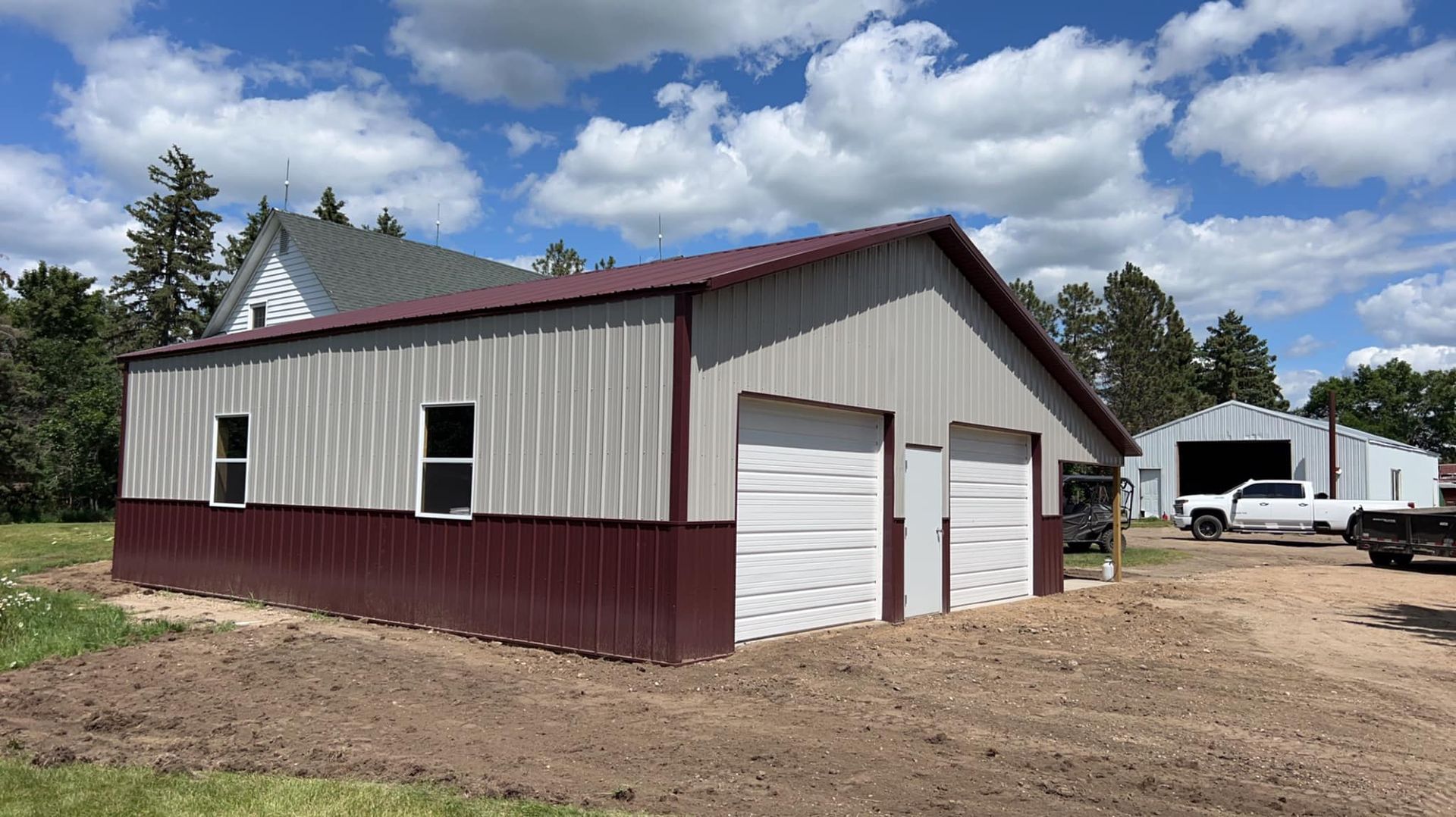 Metal garage with burgundy lower half, two garage doors, and a farmhouse in the background.