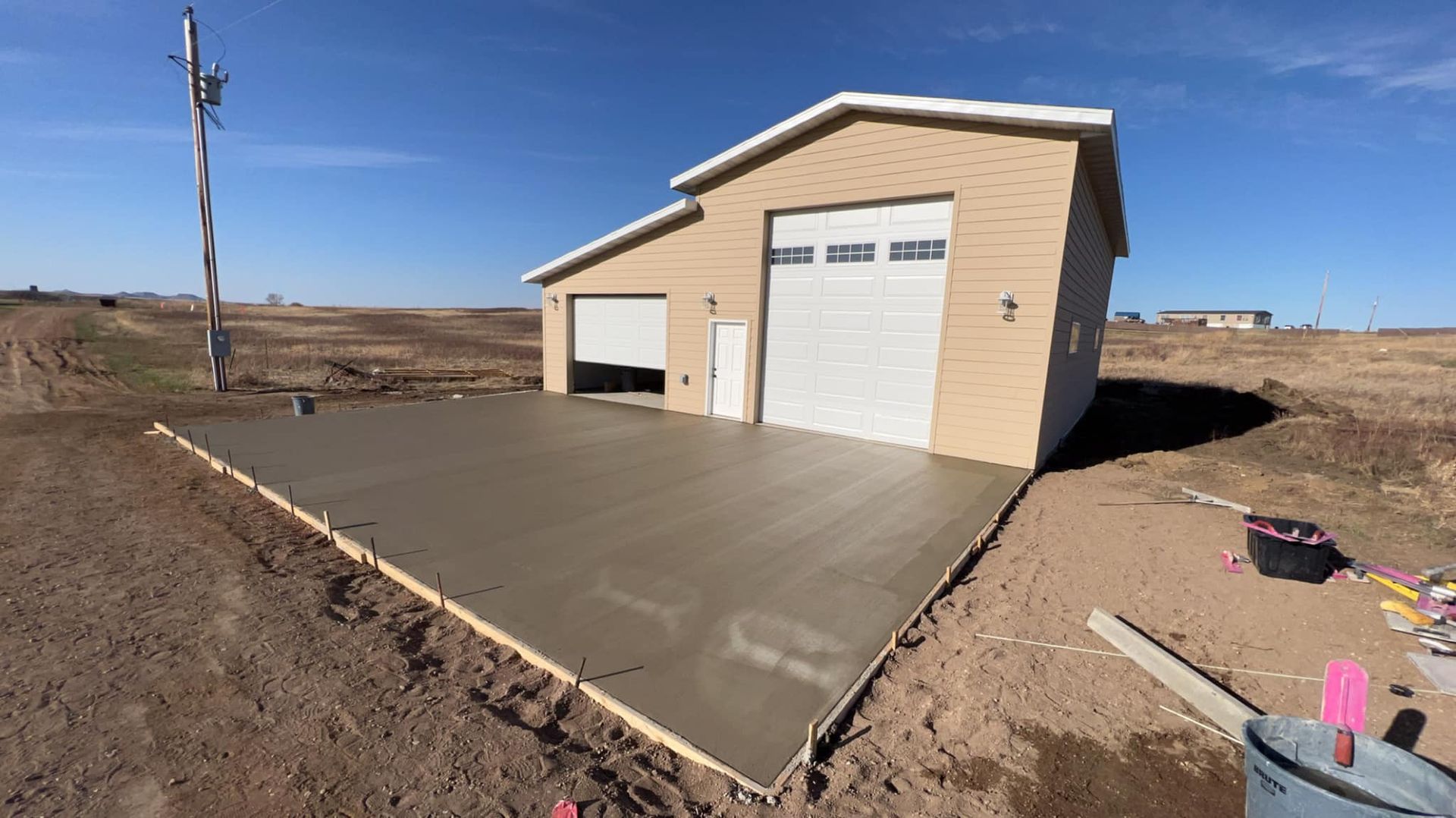 New concrete slab in front of a tan garage with two garage doors and a small door, set on brown dirt under a blue sky.