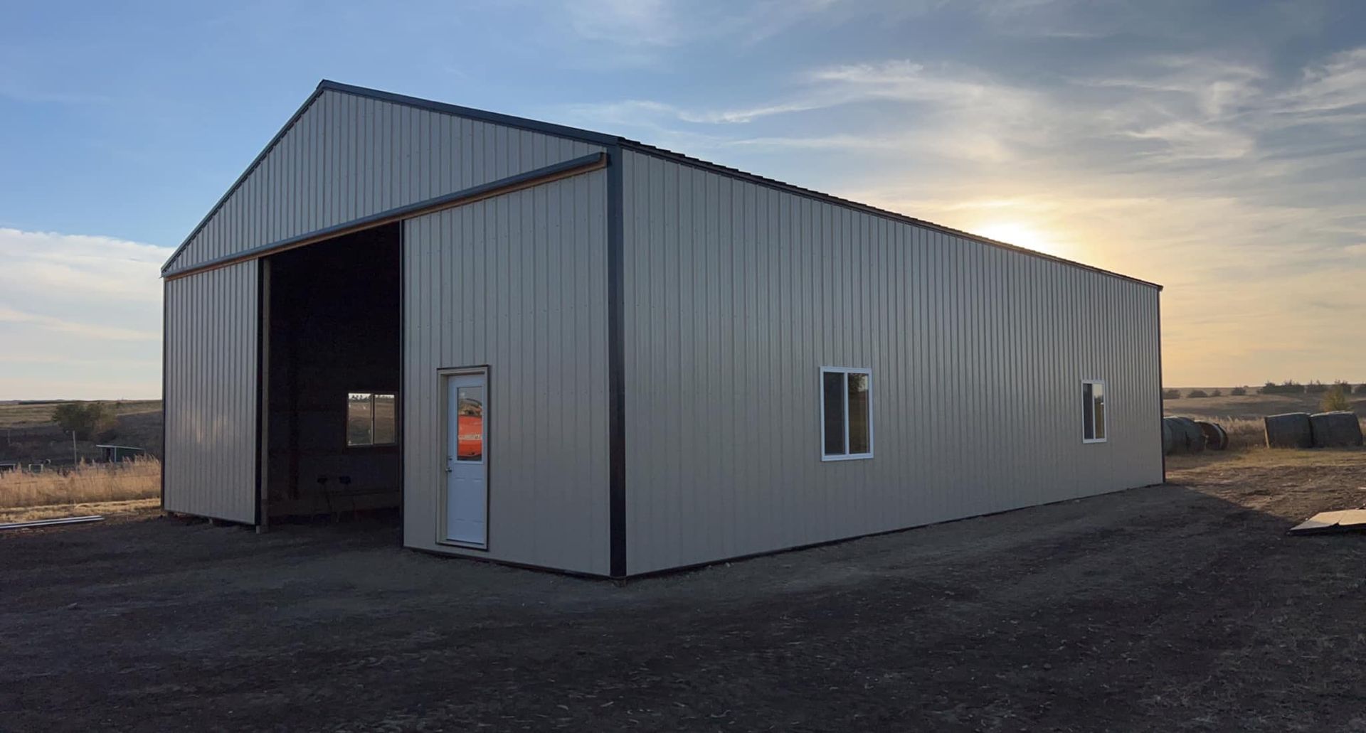 Gray metal barn with open sliding door and windows on a gravel lot under a sunny sky.