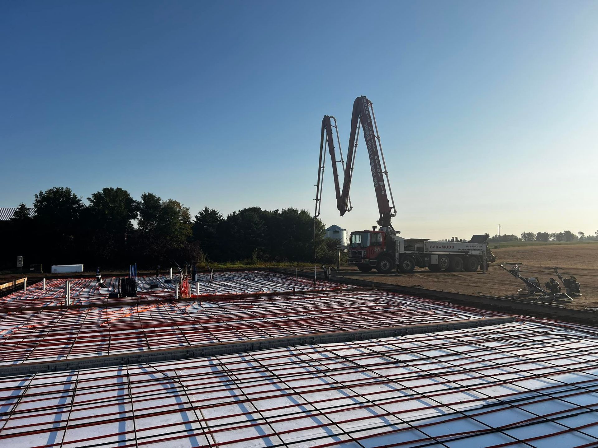 Concrete pouring at a construction site; red tubing, rebar, and concrete pump visible under a clear blue sky.
