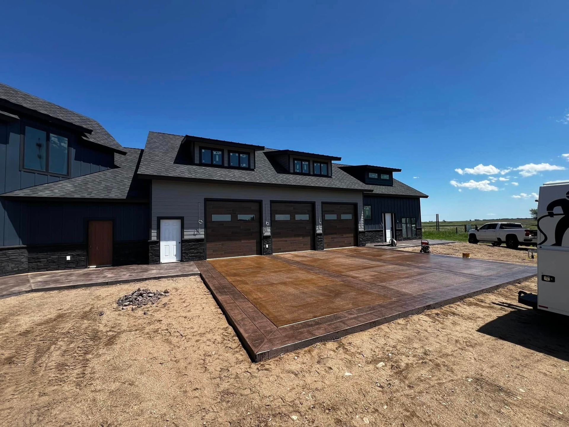 House exterior with three-car garage, brown stamped concrete patio. Blue siding, dark roof, sunny day.