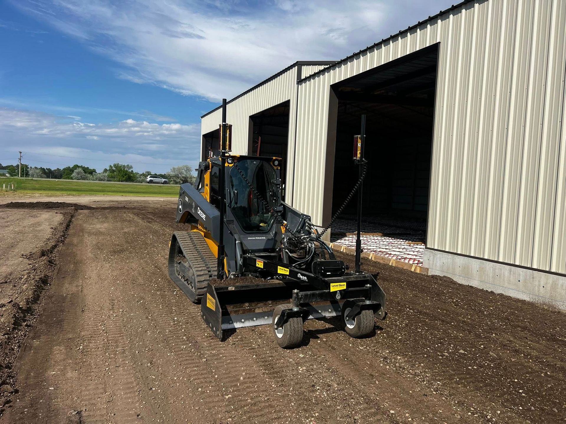 Skid steer grading gravel near a building with open garage doors; blue sky.