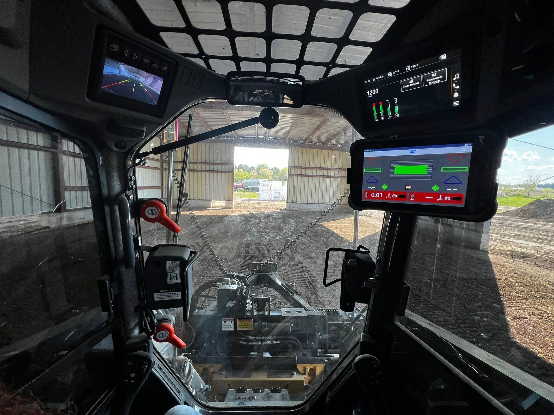 Interior view from a skid steer cab, looking out to a barn. Multiple screens show data and a rear view camera.