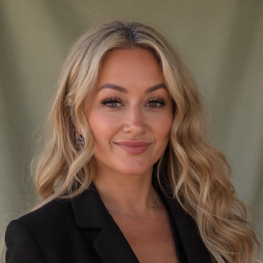 Woman with long blonde hair, smiling, wearing a black top and gray blazer.