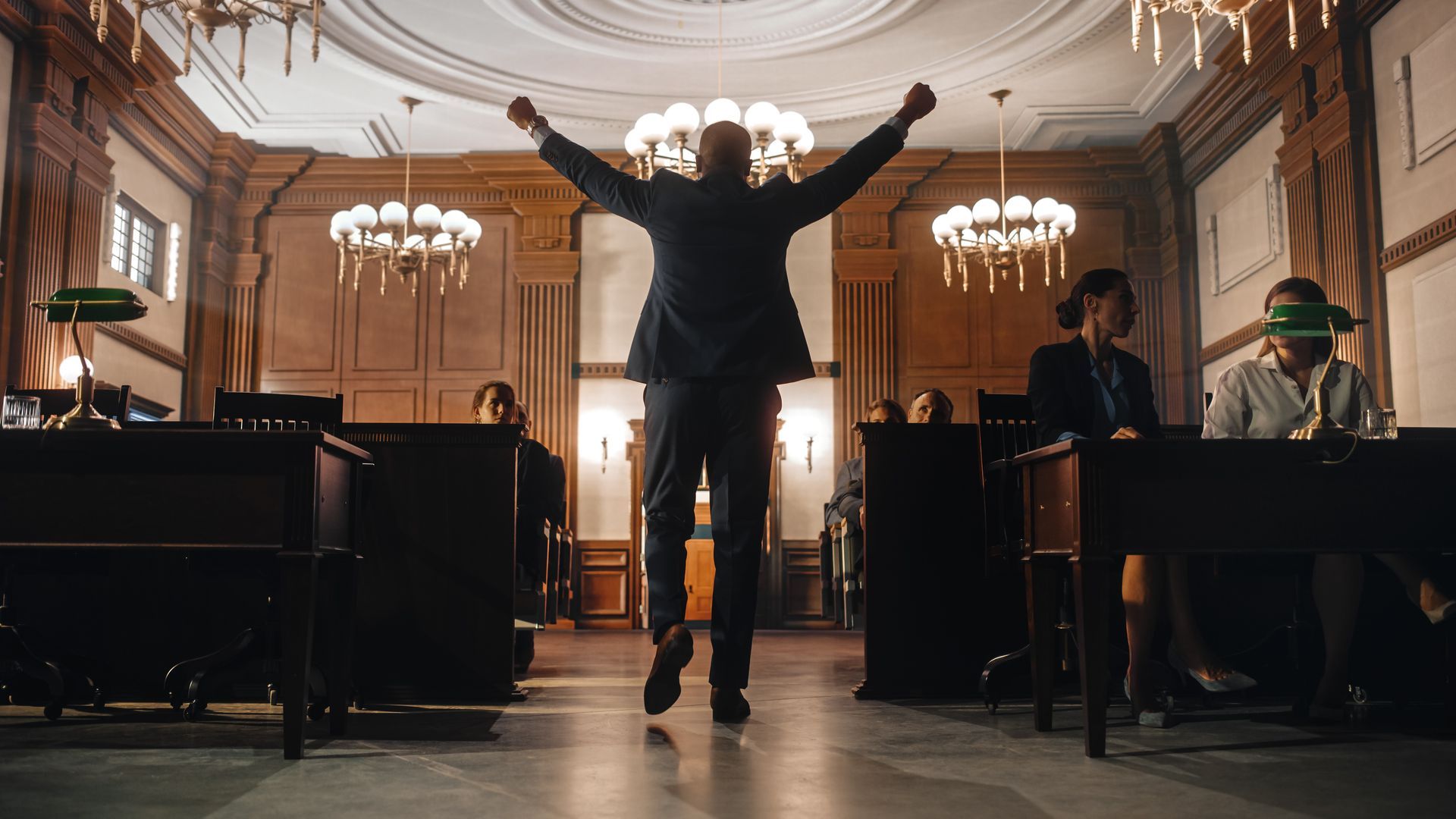 Man with arms raised celebrates in a courtroom. Other people sit at desks. Ornate room, chandeliers.