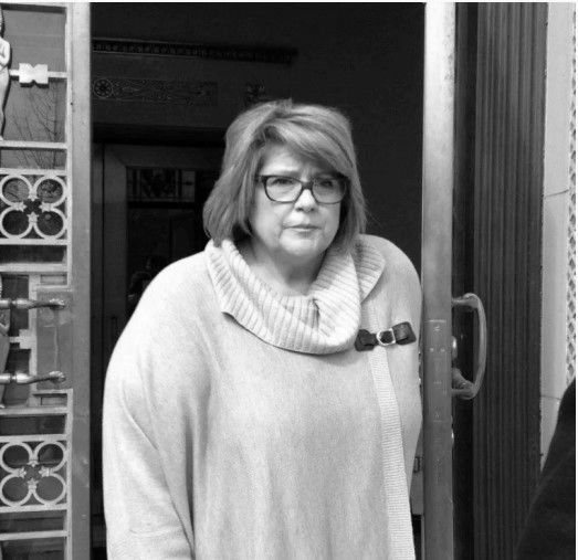 Woman with glasses, exiting a building, wearing a light-colored sweater and scarf. Black and white.