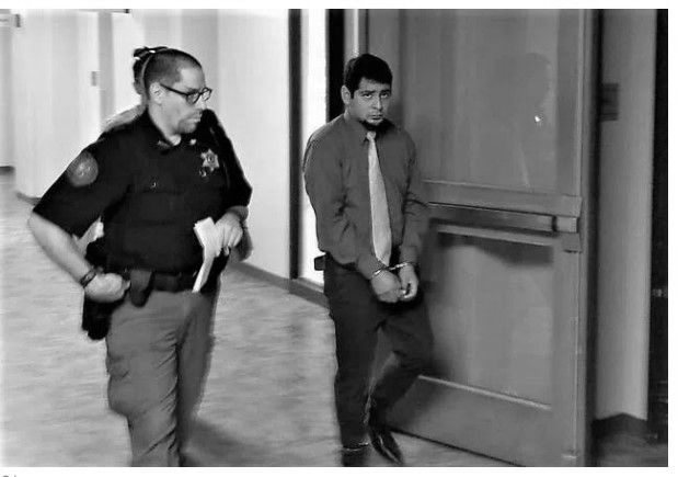 A man in handcuffs escorted by a police officer through a hallway toward a door.