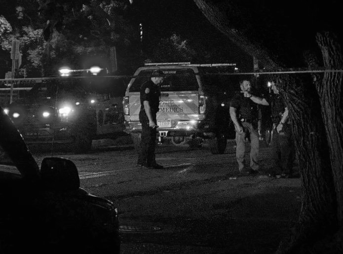 Police officers at a crime scene at night, cordoned off with tape, near vehicles and trees.