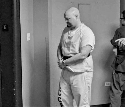 Bald man in a jail jumpsuit with hands cuffed, walking down a hallway. Another person is beside him.