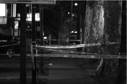 Nighttime scene with police tape cordoning off a sidewalk next to trees and a street.