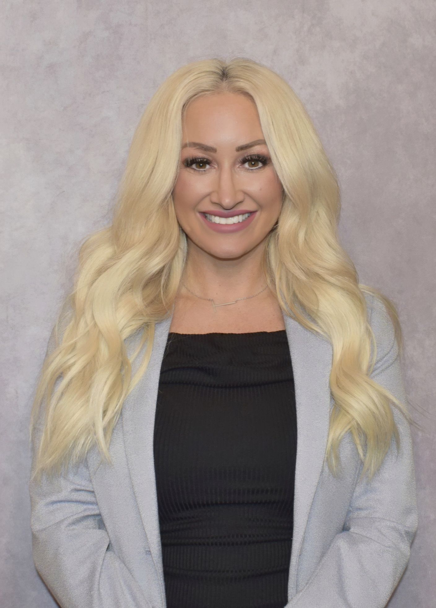 Woman with long blonde hair, smiling, wearing a black top and gray blazer.