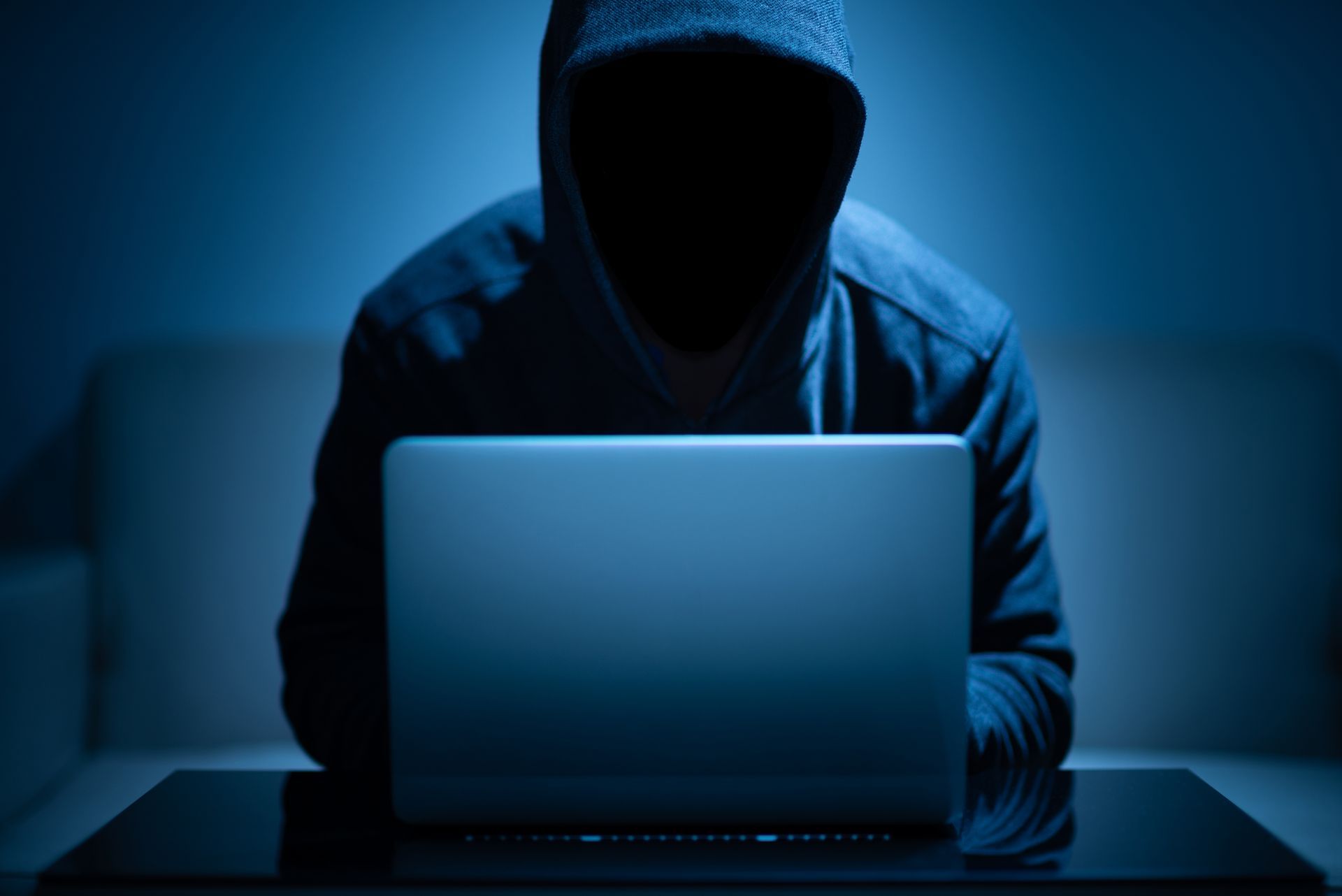 Person in a dark hoodie using a laptop in a dimly lit room.