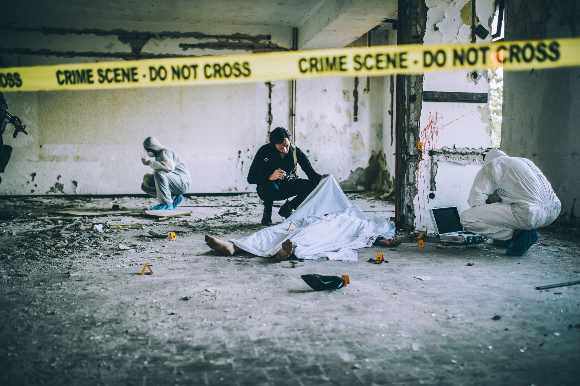 Crime scene: investigators examine a body covered by a sheet in a dilapidated building, yellow tape.