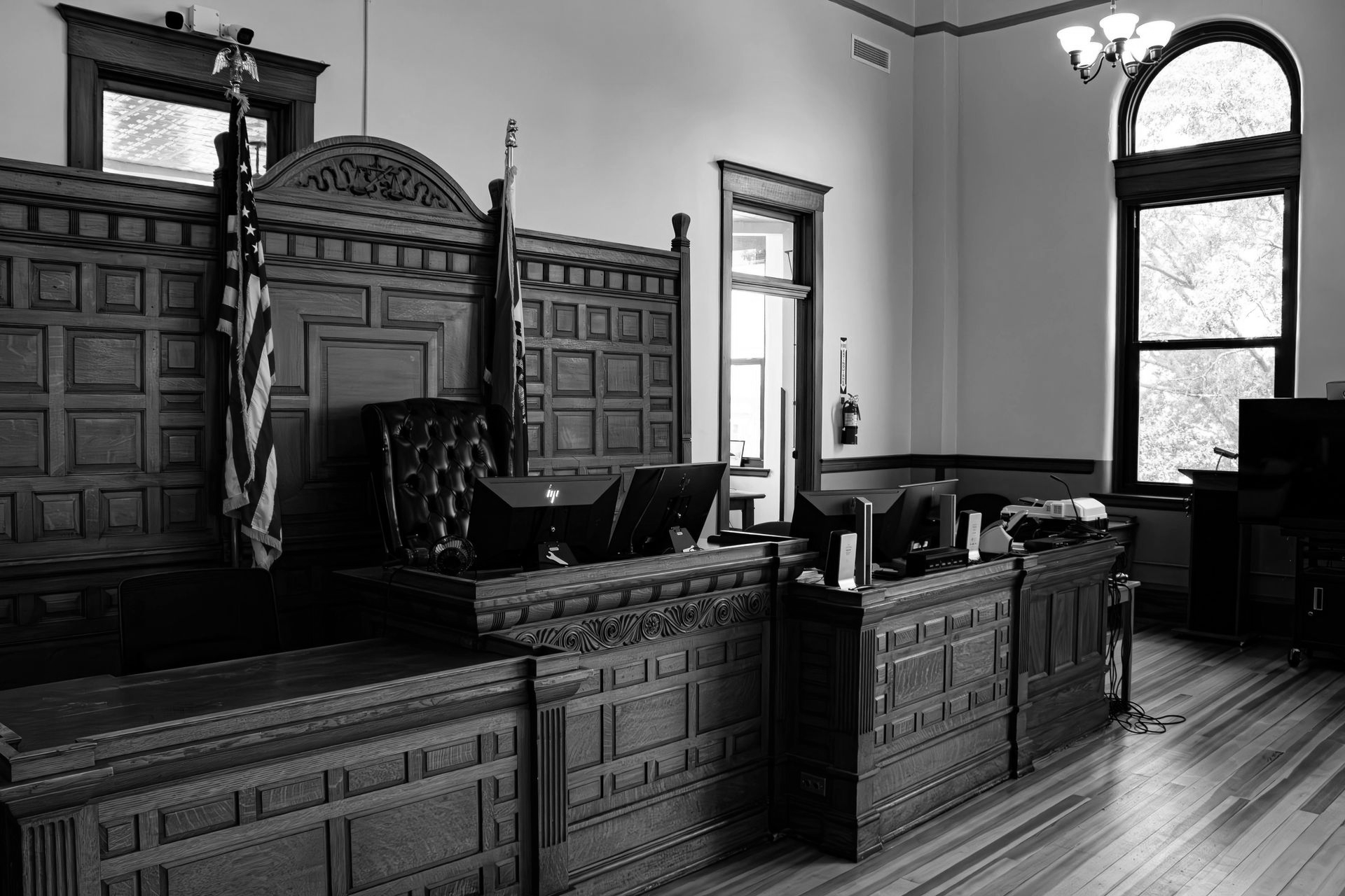 Black and white courtroom interior with wooden judge's bench, American flag, and large windows.