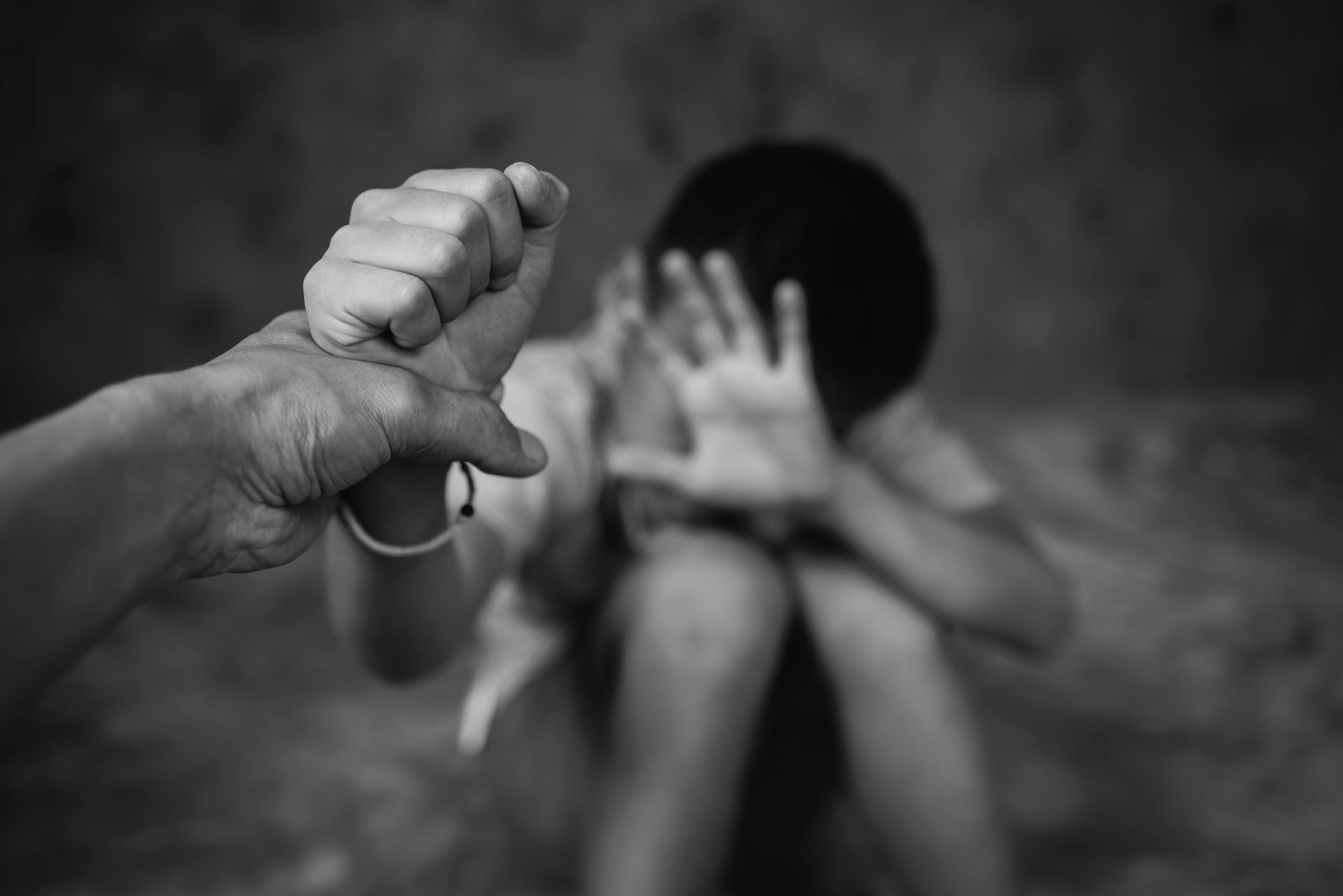 Person's fist raised towards a child covering their face, suggesting potential abuse. Black and white.