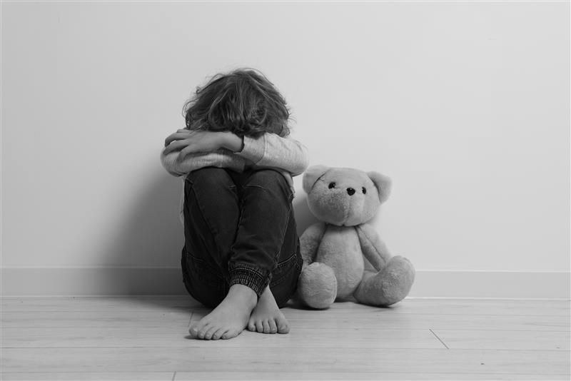 Child sitting on the floor, head in arms, next to a teddy bear.