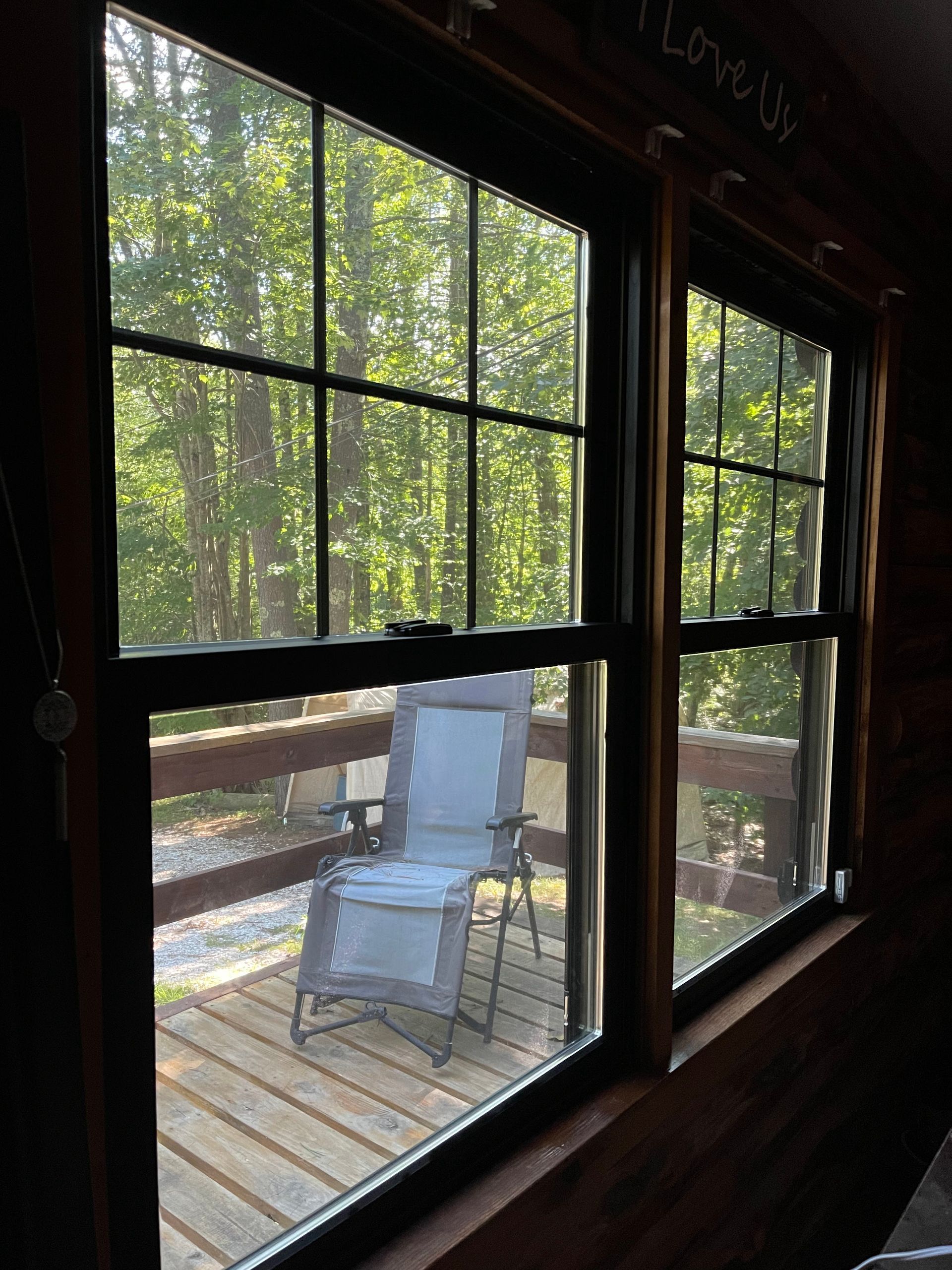 Two black-framed windows look out onto a wooden deck with a white chair and a green forest backdrop.