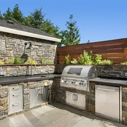 Outdoor kitchen with stainless steel appliances, stone counters and cabinetry, wooden fence and trees.
