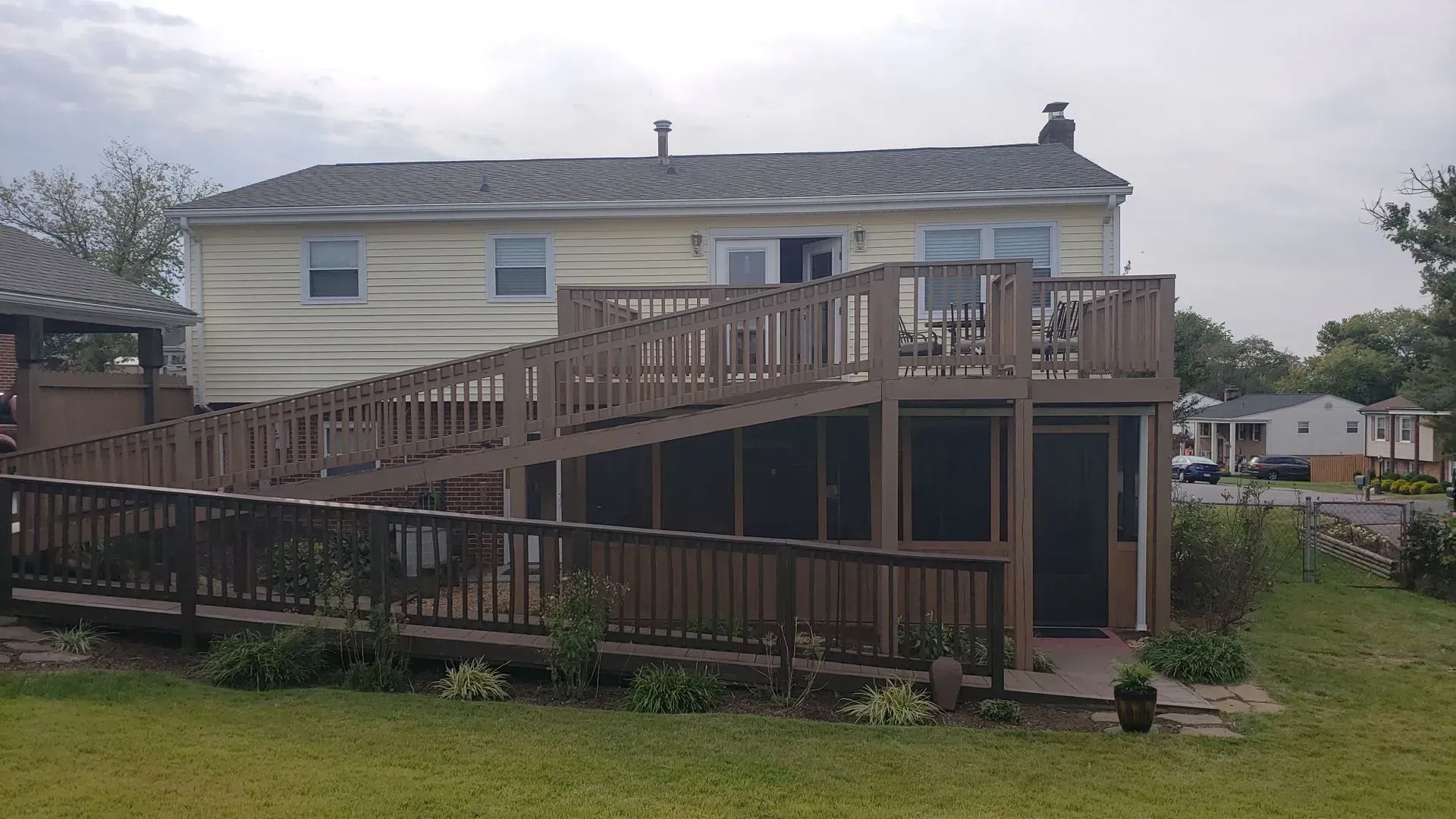 Back of a yellow house with a large wooden deck and ramp, on a grassy lawn under a cloudy sky.