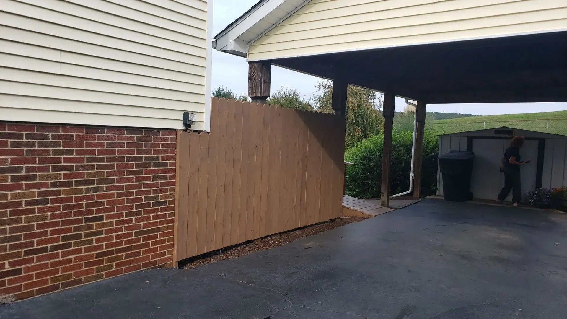 Brown wooden fence next to a brick building and carport with a person walking by.