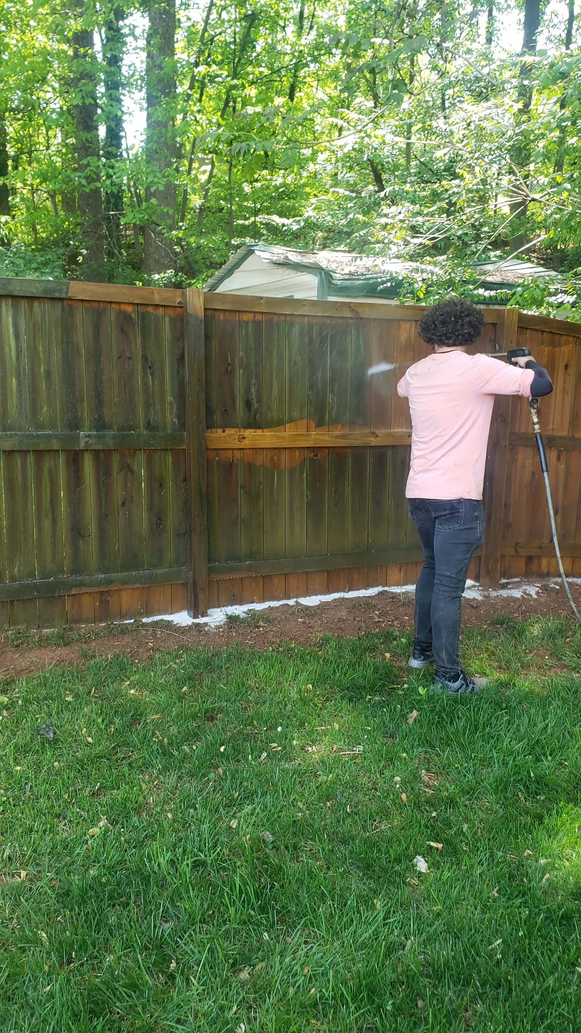 Person power washing a wooden fence in a backyard with green grass and trees.