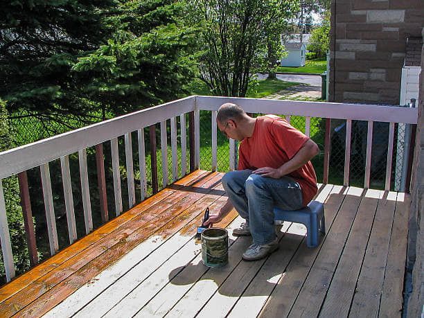 Man painting wooden deck brown, sitting on a blue stool. Bright sunlight.