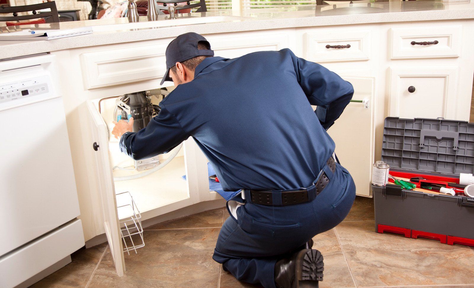 A plumber in blue uniform kneels, working on pipes under a kitchen sink. Tool box nearby.