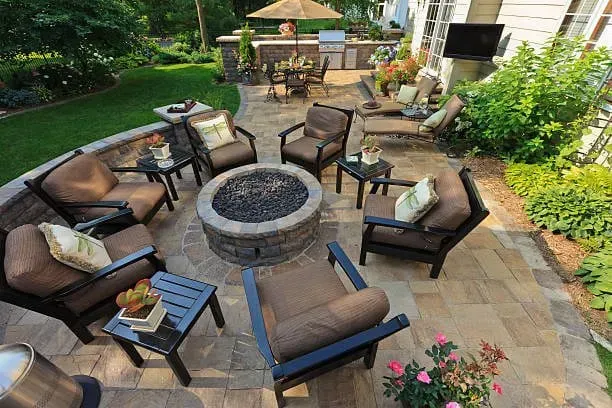 Patio with fire pit, seating area, outdoor kitchen, and green lawn. Brown chairs with beige cushions.