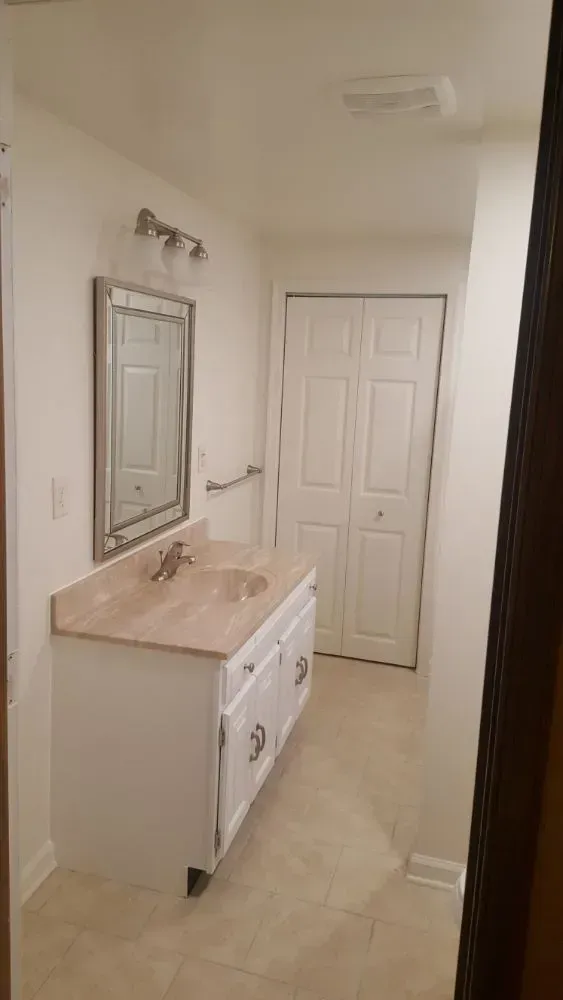 Bathroom with white vanity, mirror, and closed closet doors. Beige countertop and floor.