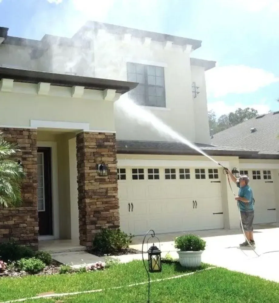Man pressure washing a two-story beige house with stone accents and a white garage door; a grassy lawn is in the foreground.