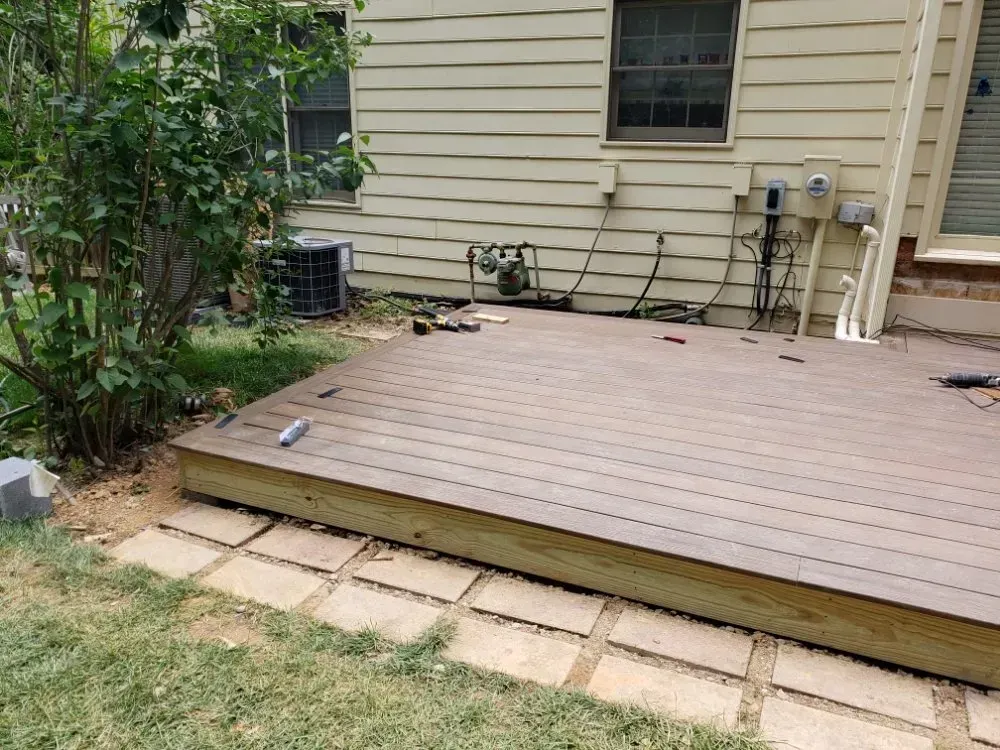 A wooden deck under construction next to a beige house. Green grass and square pavers are in the foreground.