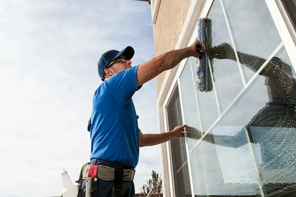 Man in blue shirt cleans a window with a squeegee, outdoors, sunny day.