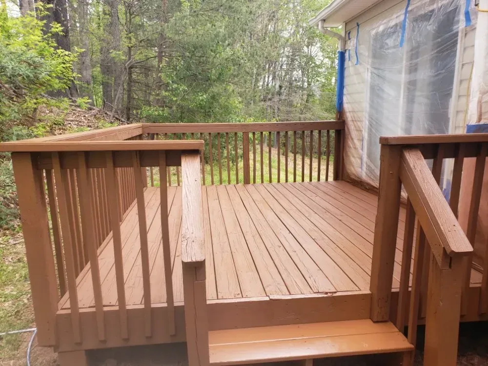 Brown wooden deck with railings, steps, and partially painted house, surrounded by trees.