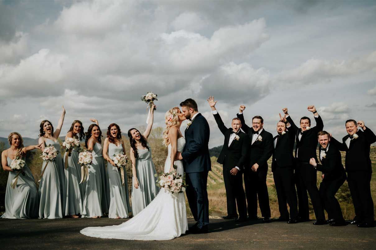 A bride and groom are kissing in front of their wedding party.