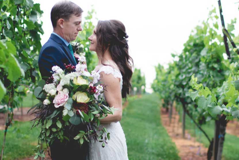 A bride and groom are standing in a vineyard holding a bouquet of flowers.