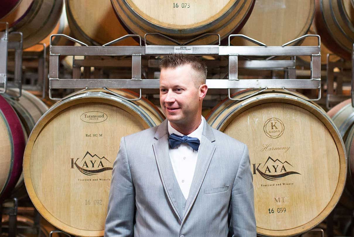 A man in a suit and bow tie is standing in front of a row of wine barrels.