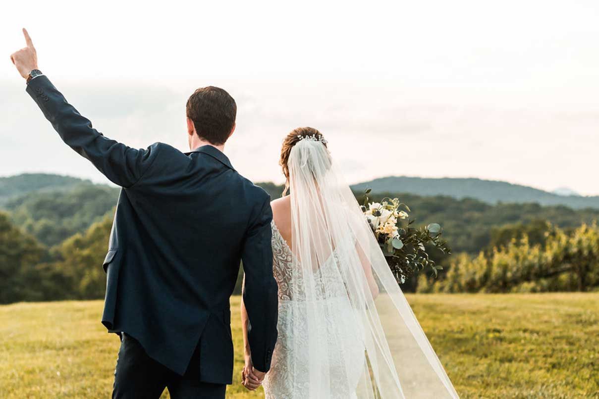 A bride and groom are walking in a field with mountains in the background.