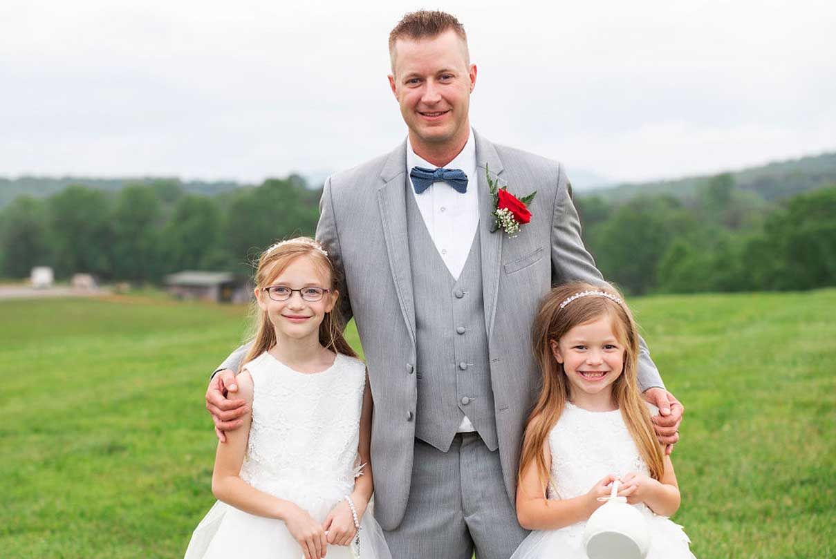 A groom and two flower girls are posing for a picture in a field.