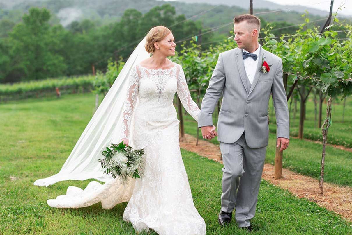 A bride and groom are walking through a vineyard holding hands.