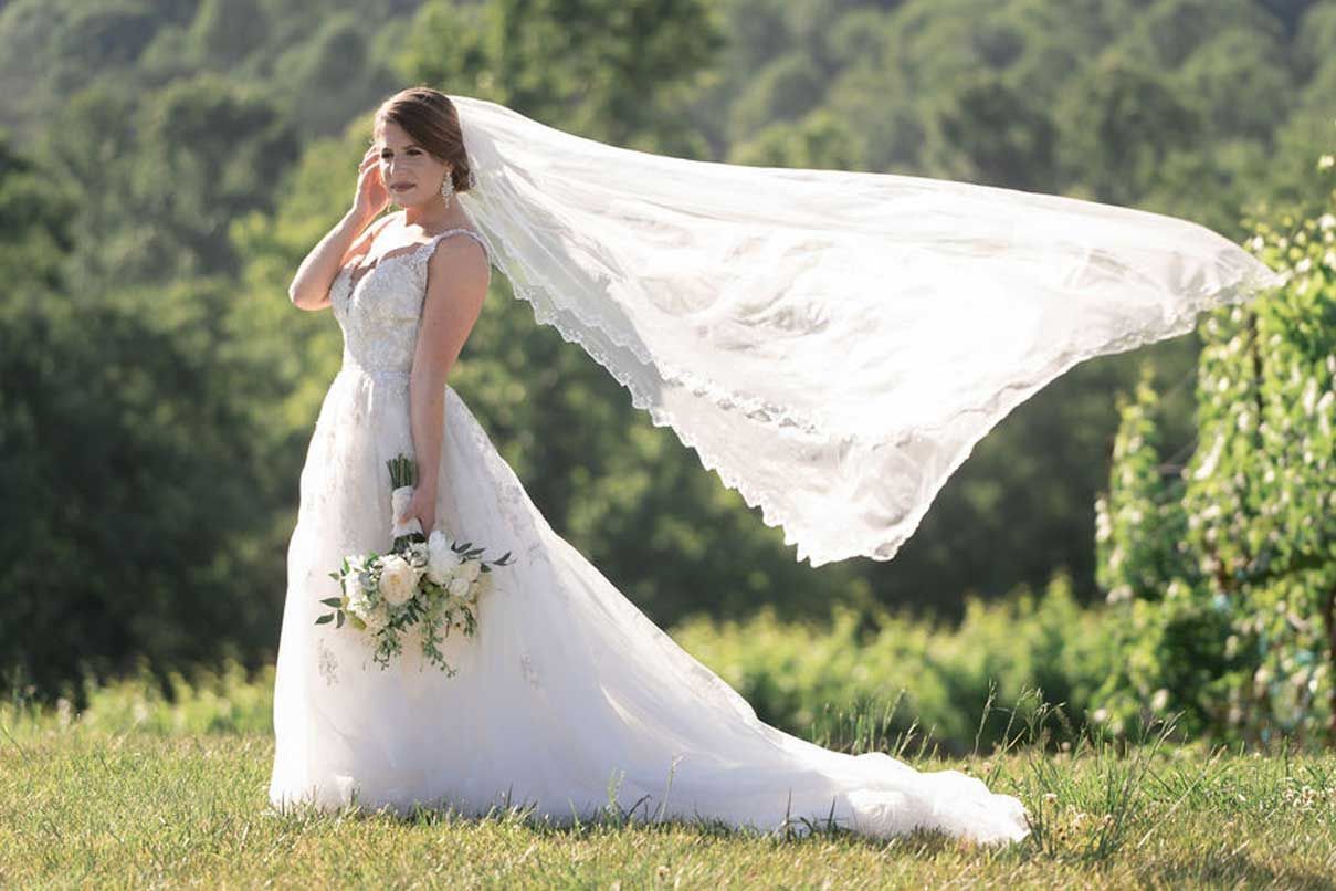 A bride in a wedding dress is standing in a field with her veil blowing in the wind.