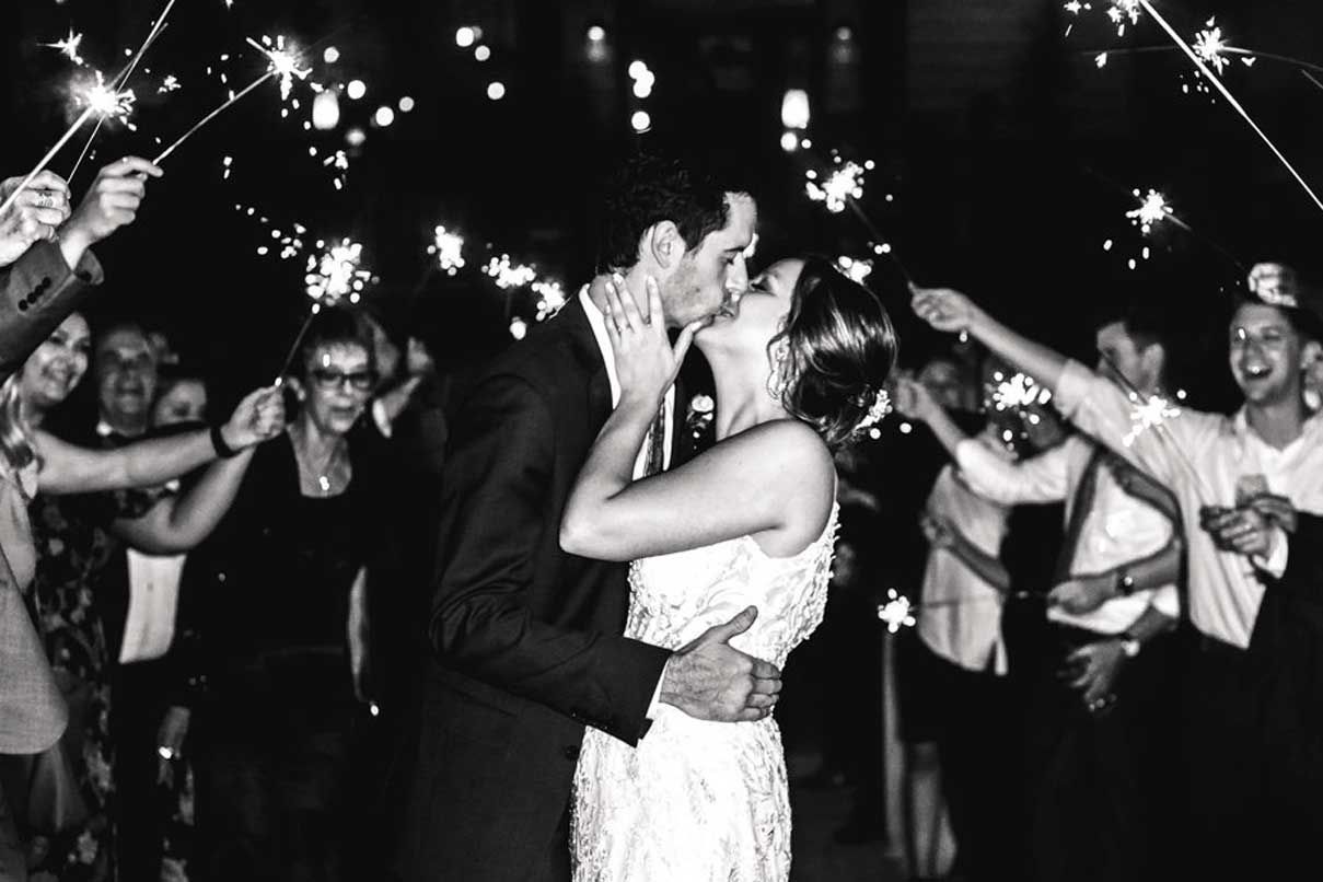 A bride and groom kissing in front of a crowd of people holding sparklers.
