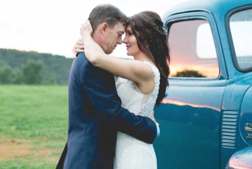 A bride and groom are hugging in front of a blue truck.