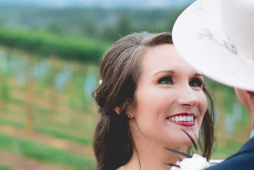 A bride and groom are looking at each other in a vineyard . the bride is wearing a cowboy hat.