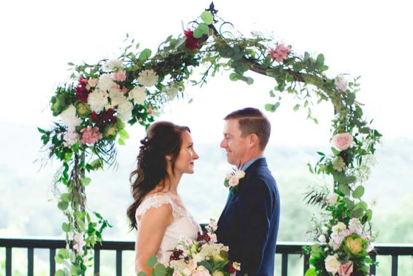 A bride and groom are standing under a floral arch at their wedding.