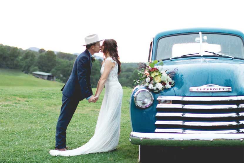 A bride and groom are kissing in front of a blue truck.