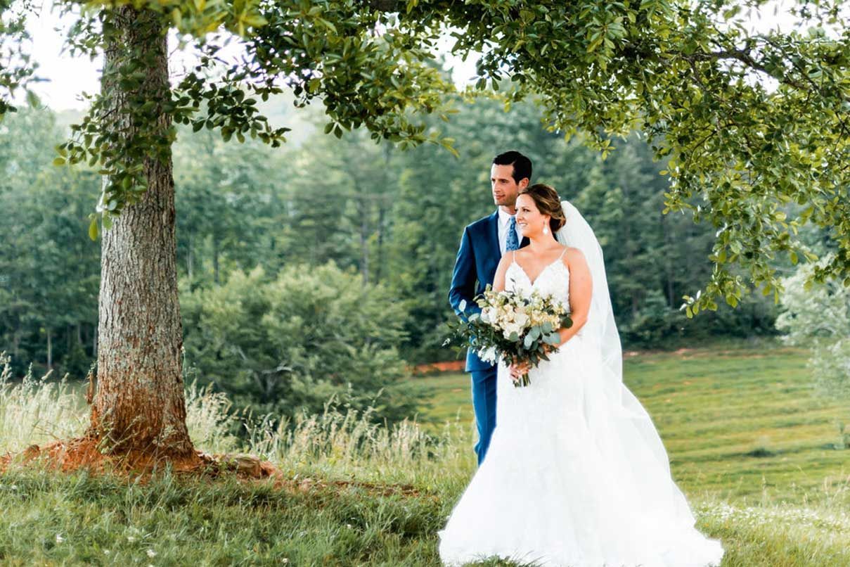 A bride and groom are standing under a tree in a field.