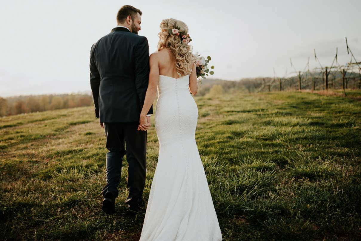 A bride and groom are standing in a field holding hands.