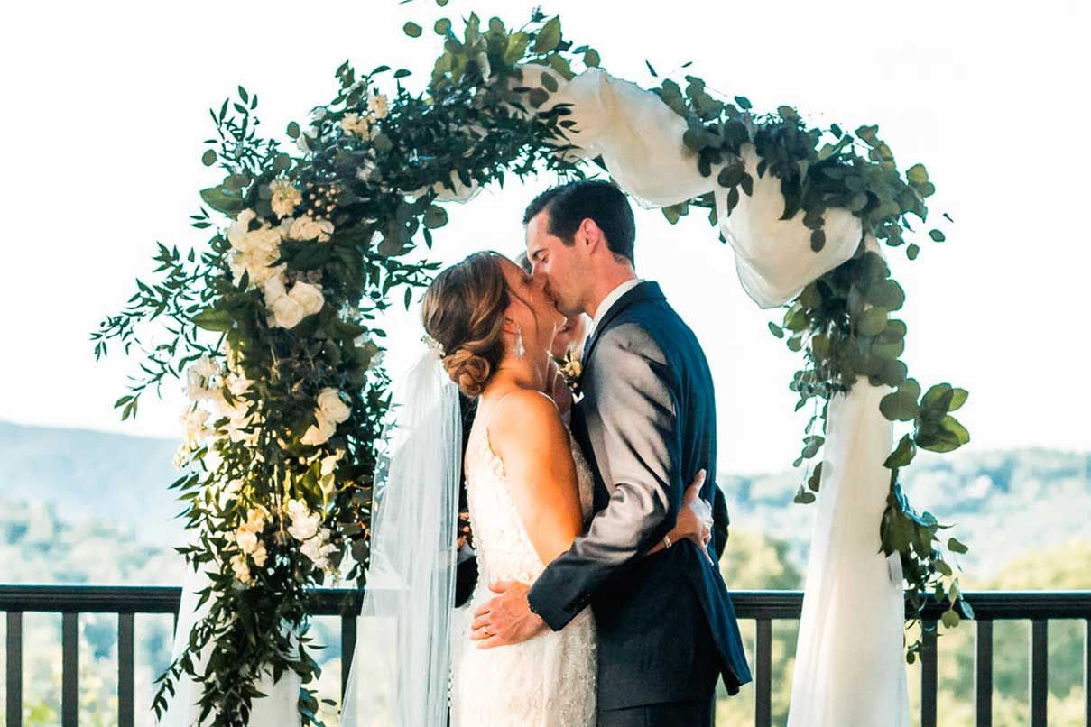 A bride and groom are kissing under a floral arch at their wedding.
